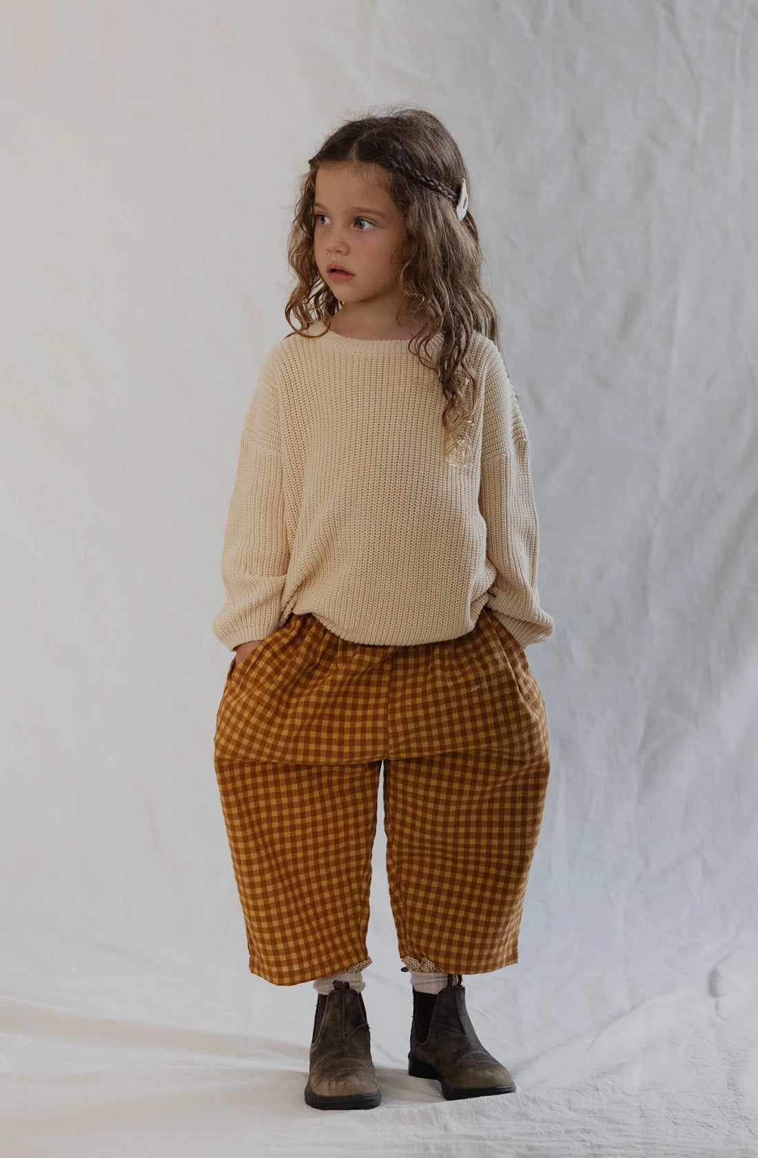 A young child with long curly hair stands against a white fabric backdrop, wearing a beige sweater, VALENCIA BYRON BAY Rio Pant Honeycomb in brown and yellow checkered patterns, and brown ankle boots. With their hands in their pockets, the unisex design of the outfit complements the serious expression on their face.