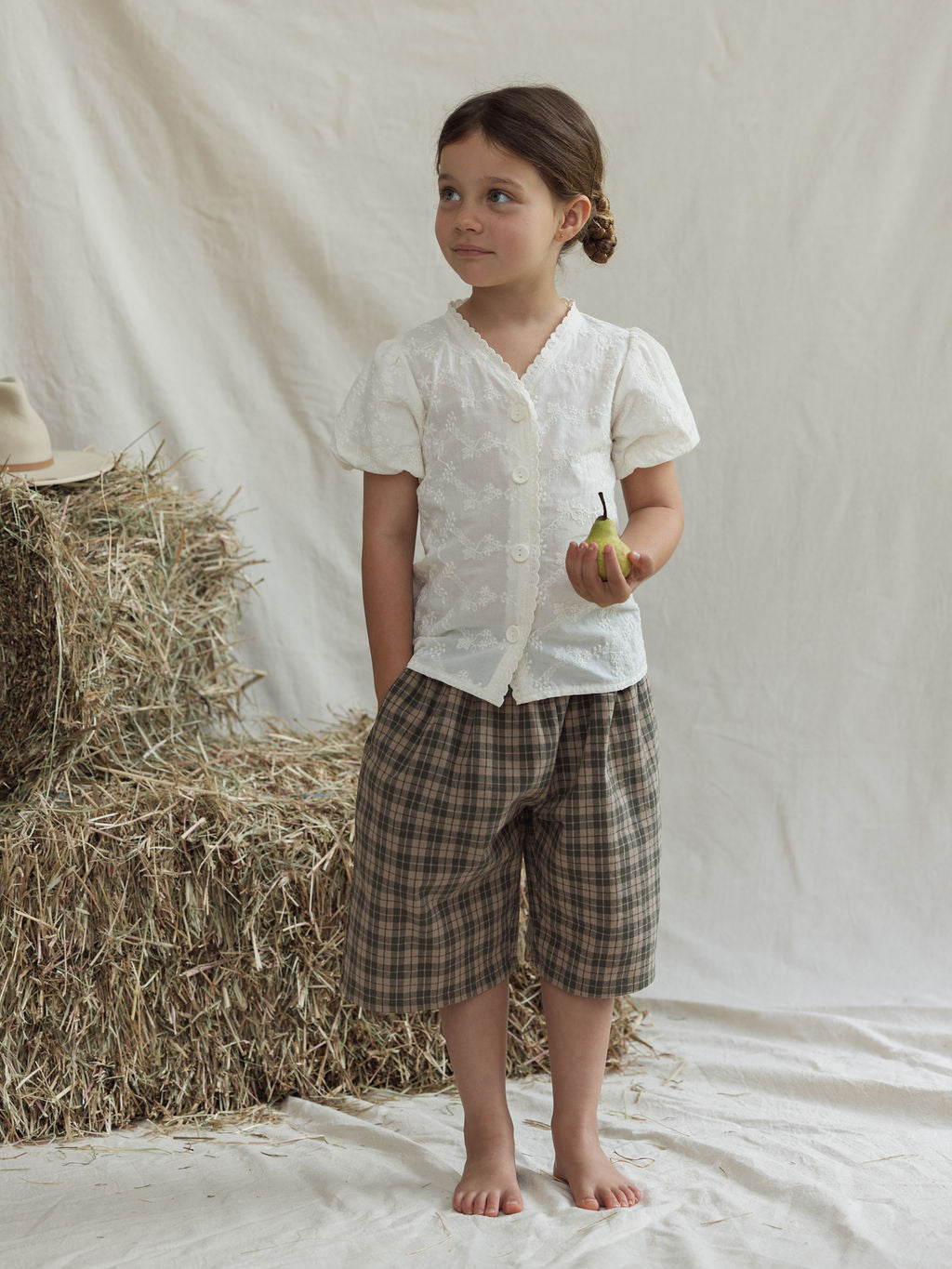 A young girl stands barefoot on white fabric in front of a hay bale. She is wearing the Hinterland Blouse by VALENCIA BYRON BAY and plaid shorts, holding a pear in her right hand. Her expression is neutral, with a hat resting on the hay bale.