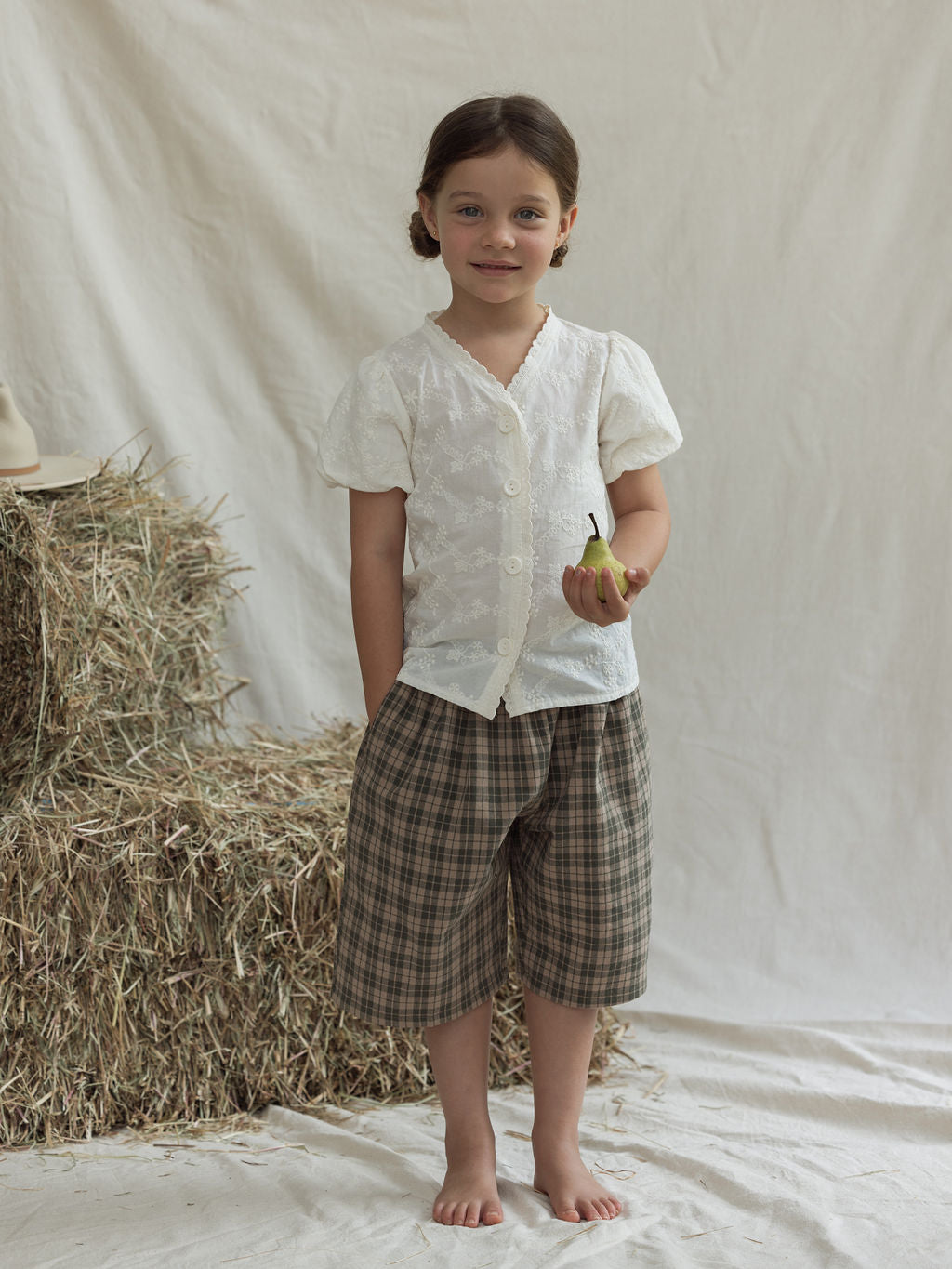 A child stands barefoot on a white sheet, wearing the Hinterland Blouse from VALENCIA BYRON BAY, featuring floral embroidered details, paired with plaid shorts and holding a small green apple. Behind the child is a stack of hay bales on the left.