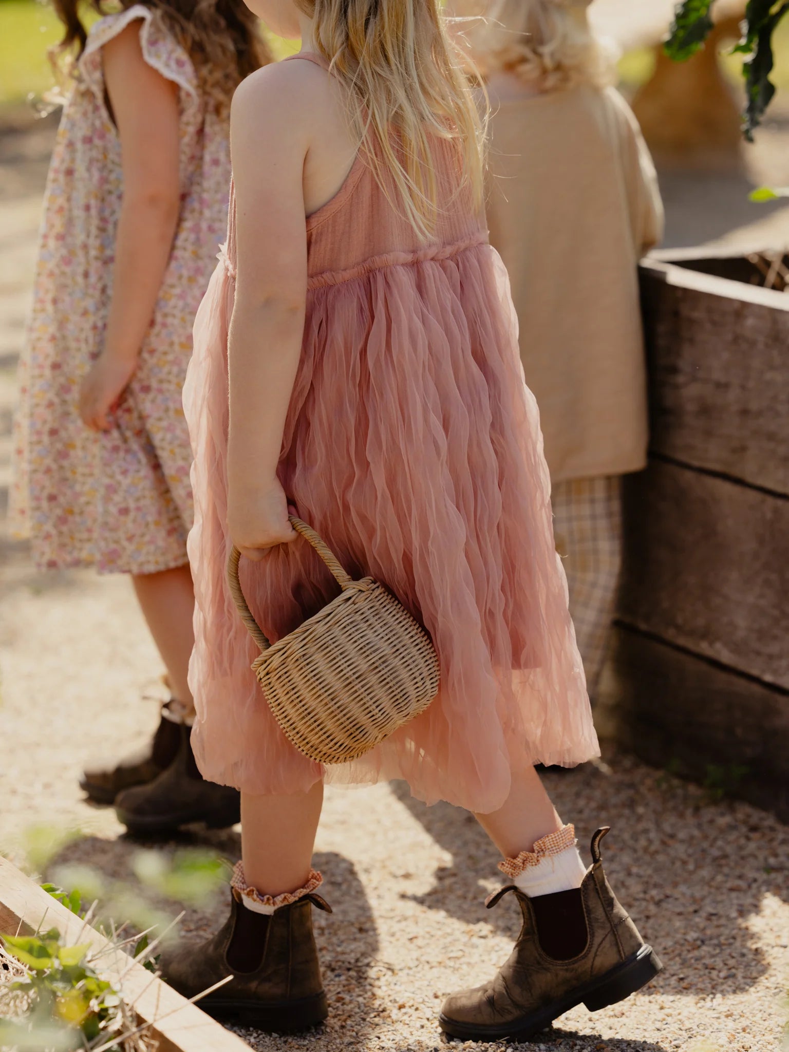 Three children are walking outside near a wooden planter box. They are wearing light, summery clothing; one child, in the foreground, is dressed in a Bangalow Tutu Dress Antique Rose by VALENCIA BYRON BAY and holds a small, woven basket. The background shows a sandy path and blurred greenery.