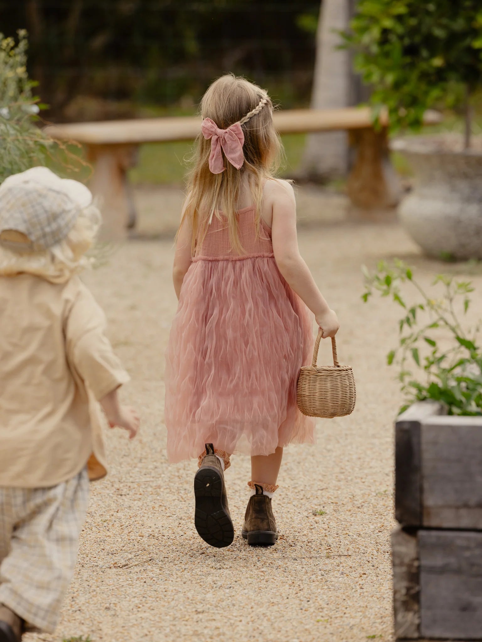 A young girl with long hair, adorned with a floral headband and a pink bow, walks away on a gravel path. She is wearing the VALENCIA BYRON BAY Bangalow Tutu Dress in Antique Rose and brown boots, carrying a small woven basket. A younger child in a beige outfit and checkered cap follows her. Greenery surrounds the path.