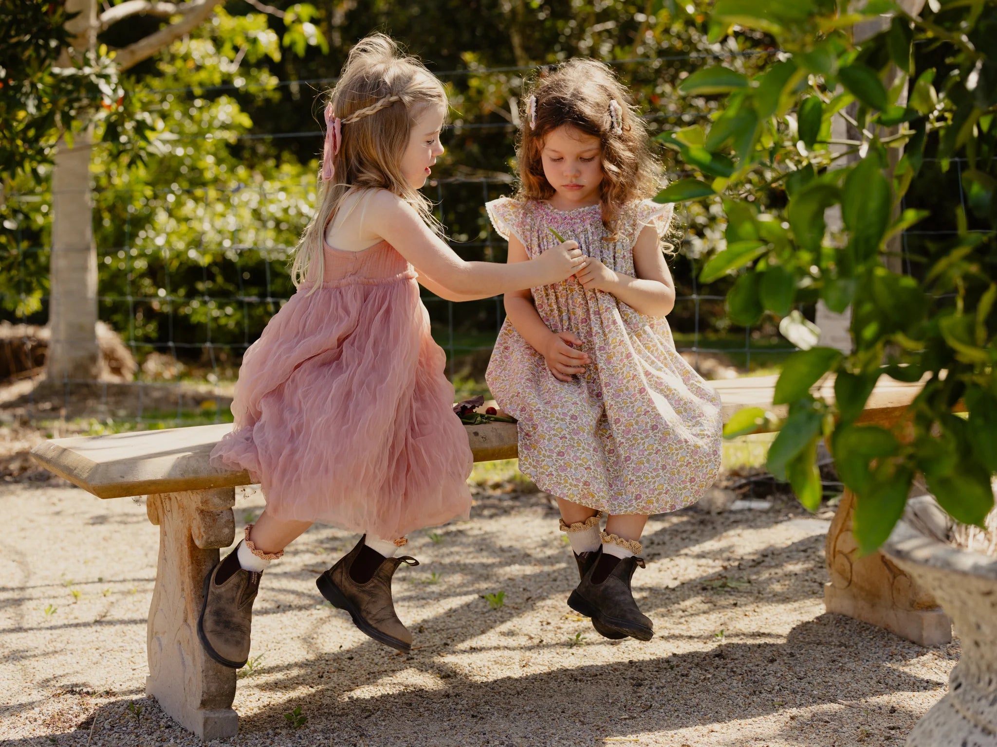 Two young girls are sitting on a wooden bench in a sunny garden. The girl on the left, wearing the Bangalow Tutu Dress in Antique Rose by Valencia Byron Bay and brown boots, is handing something to the girl on the right, who is dressed in a floral cotton muslin dress and brown boots. The background is filled with greenery.