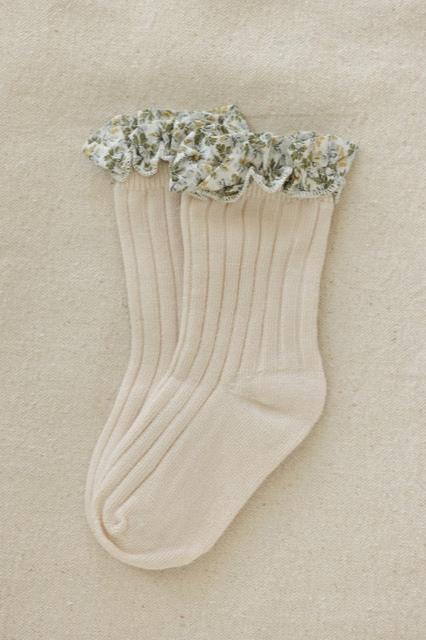 A pair of sand-colored socks with a ribbed texture, featuring a Coorabell floral-printed frill at the top. The floral trim has a green and white design. The socks, part of Valencia Byron Bay's Frilly Socks collection, are neatly laid out on a beige fabric background.