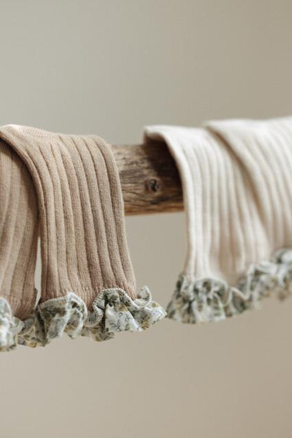 Close-up of two pairs of textured, sand-colored baby socks hanging on a wooden rod. The socks, known as Coorabell Socks Sand by Valencia Byron Bay, feature ribbed patterns and are adorned with delicate floral-printed frills at the cuffs. The background is out of focus, highlighting the intricate details of the socks.