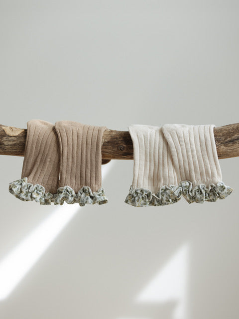 A wooden branch holds two pairs of ribbed, ruffled socks. One pair is Coorabell Socks Sand from VALENCIA BYRON BAY with floral trim, and the other is cream with a touch of Valencia's Frilly Socks. The background is softly lit with natural light, creating a minimalist, serene atmosphere.
