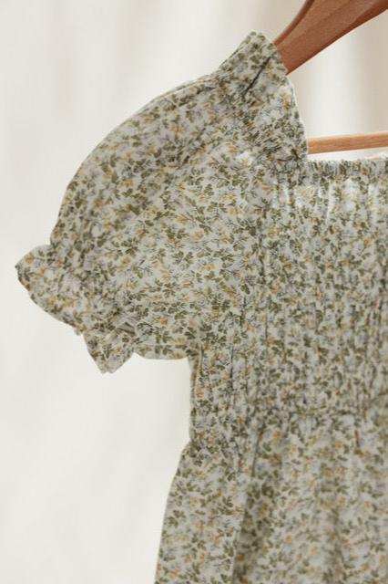 A close-up of the VALENCIA BYRON BAY Coorabell Dress showcases its short puffed sleeves and ruched bodice. Made from soft, breathable cotton, the fabric boasts a delicate pattern of green leaves and small yellow flowers. This feminine dress is displayed on a wooden hanger against a light background.