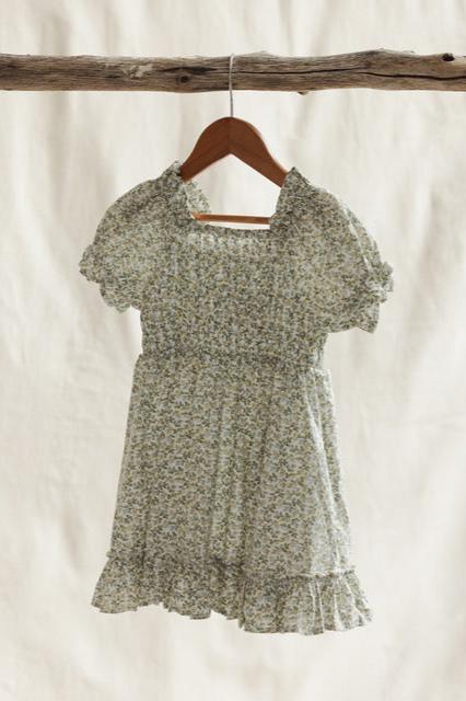 A light green floral Coorabell Dress by VALENCIA BYRON BAY, made from soft, breathable cotton with short puffed sleeves, hangs on a wooden hanger against a beige background. The feminine design features a gathered bodice and a ruffled hemline.