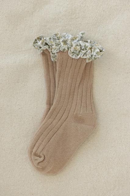 A pair of Coorabell Socks Oat, featuring a beige ribbed design with a floral-patterned ruffle at the top. These socks, part of VALENCIA BYRON BAY's Frilly Socks collection, are laid flat on a light-textured fabric surface.