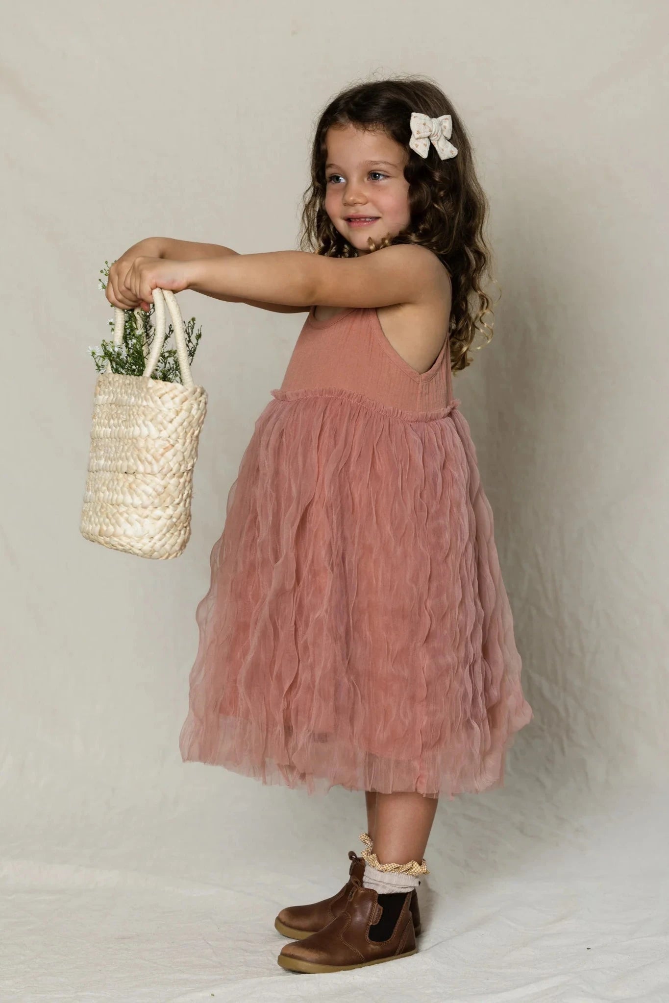 A young girl with curly hair wears a pink tulle "Bangalow Tutu Dress Antique Rose" by VALENCIA BYRON BAY and brown boots with socks. She stands sideways, smiling, and holds a small white basket filled with greenery. A bow adorns her hair, and the backdrop is a plain off-white color.