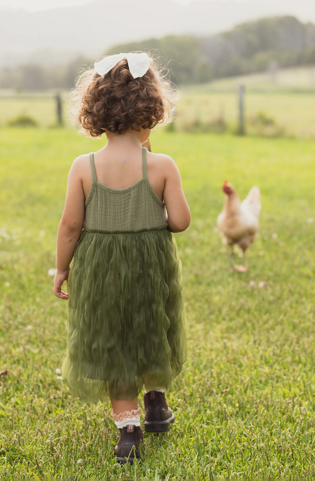 A young child with curly hair, wearing a green Bangalow Tutu Dress Eucalyptus from VALENCIA BYRON BAY and a white bow on their head, stands on a green field, looking at a chicken in the distance. The child has their back to the camera, and the scene is set in a rural, outdoor area with soft, warm light.