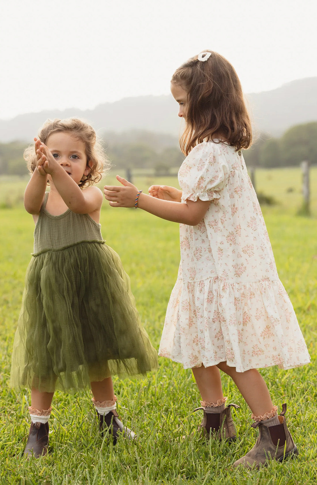 Two young children play on a grassy field. The child on the left, in a green dress, claps their hands joyfully, while the child on the right is in a white floral Bangalow Tutu Dress Eucalyptus by VALENCIA BYRON BAY, holding hands with their friend. Both have curly hair and are wearing brown boots.