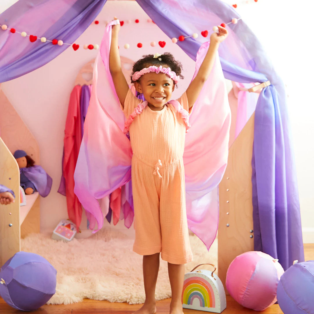 A young girl joyfully stands in a play area adorned with SARAH'S SILKS' Large Blossom Streamer. She's dressed in a peach outfit and a flower headband, her arms outstretched. Beside her, she holds a wooden wand near toys like a rainbow bag and plush balls, creating a cheerful and imaginative setting.