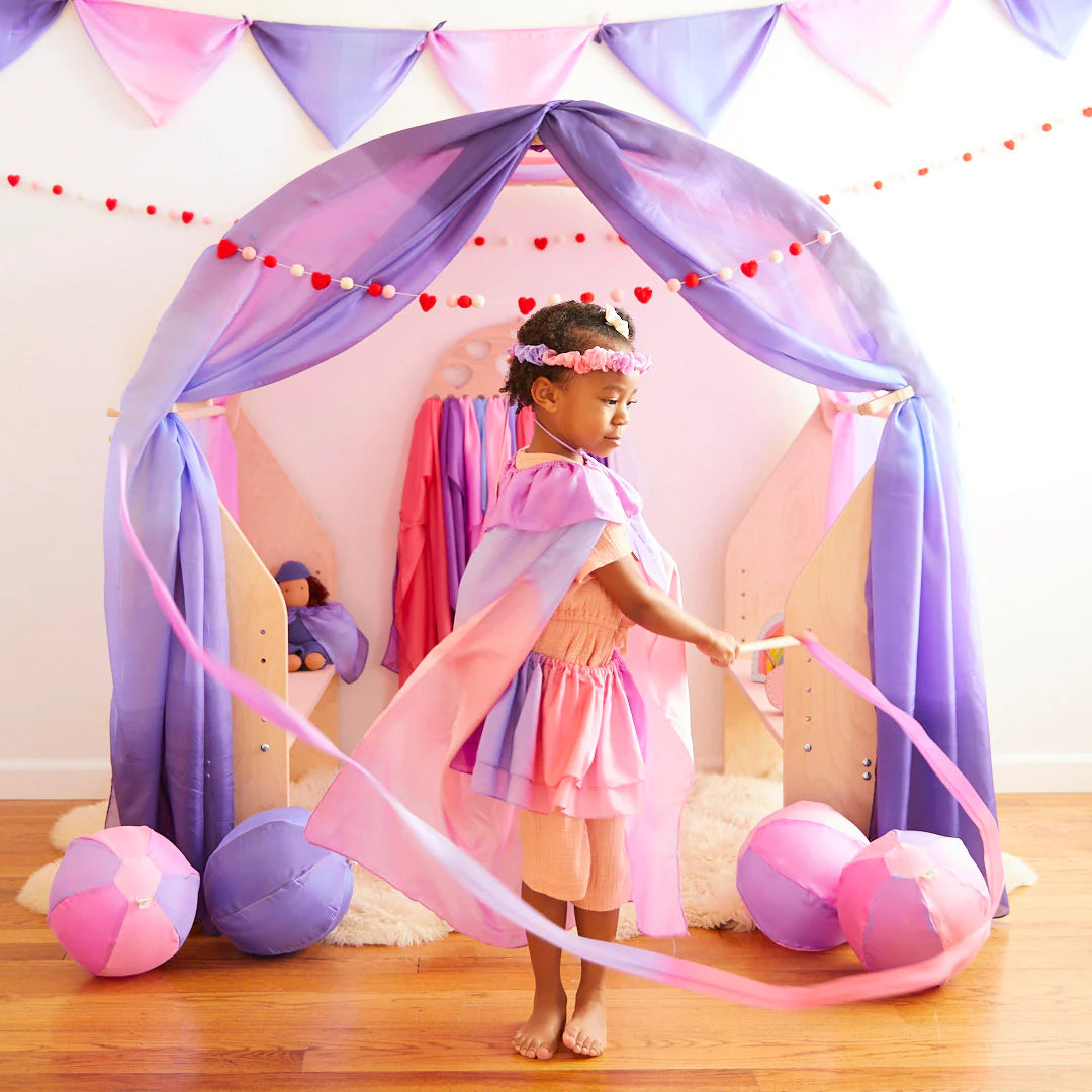 A young child dressed as a princess twirls the Large Blossom Streamer by SARAH'S SILKS with delight, surrounded by colorful ribbons in front of a pastel-colored play tent. Adorned with pink and purple fabrics, matching bunting, and plush toys on the floor, she clutches her wooden wand made from sustainably sourced materials.