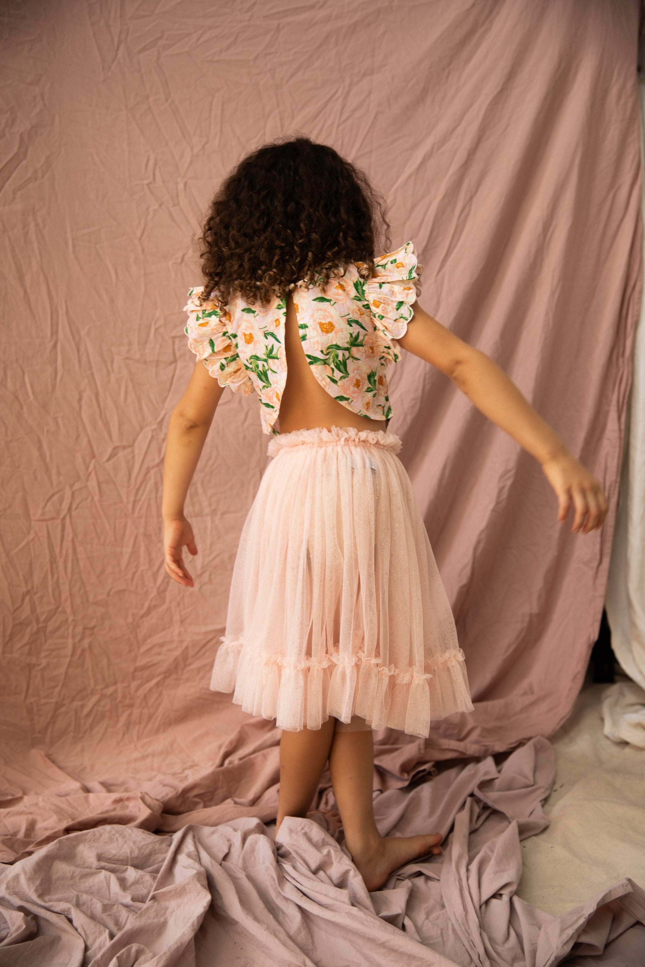 A young girl with curly hair stands on a draped fabric background, facing away from the camera. She is wearing the BELLA + LACE Vivian Top Soirée Bouquet/Strawberry Sundae, which features ruffled sleeves and an open back design. The top is made of light cotton voile and paired with a knee-length tulle skirt adorned with scalloped embroidery at the hem.