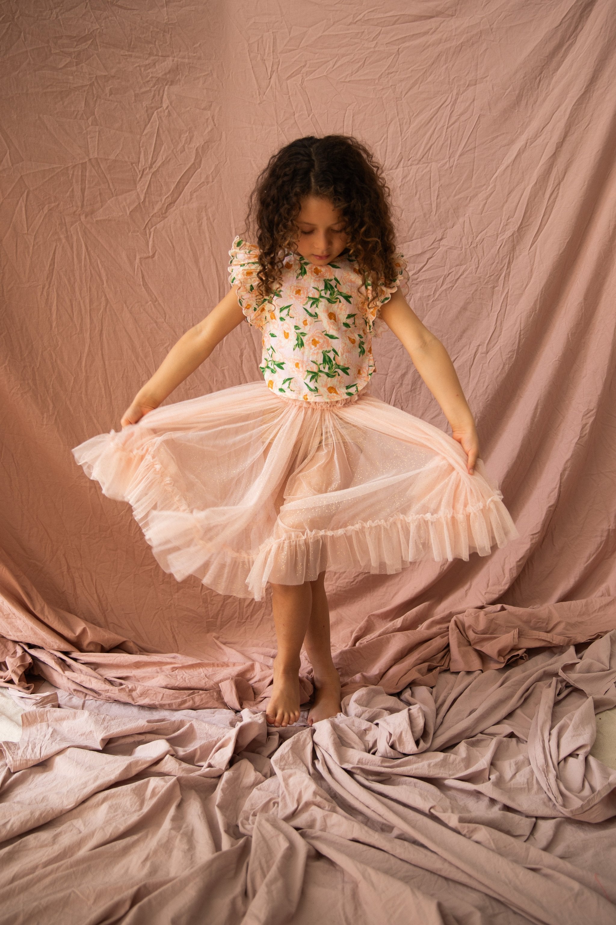 A young girl with curly hair stands barefoot on wrinkled fabric, looking down at her light pink tulle skirt, which she is holding out in both hands. She wears the BELLA + LACE Vivian Top Soirée Bouquet/Strawberry Sundae, set against a backdrop of soft, pink draped fabric.