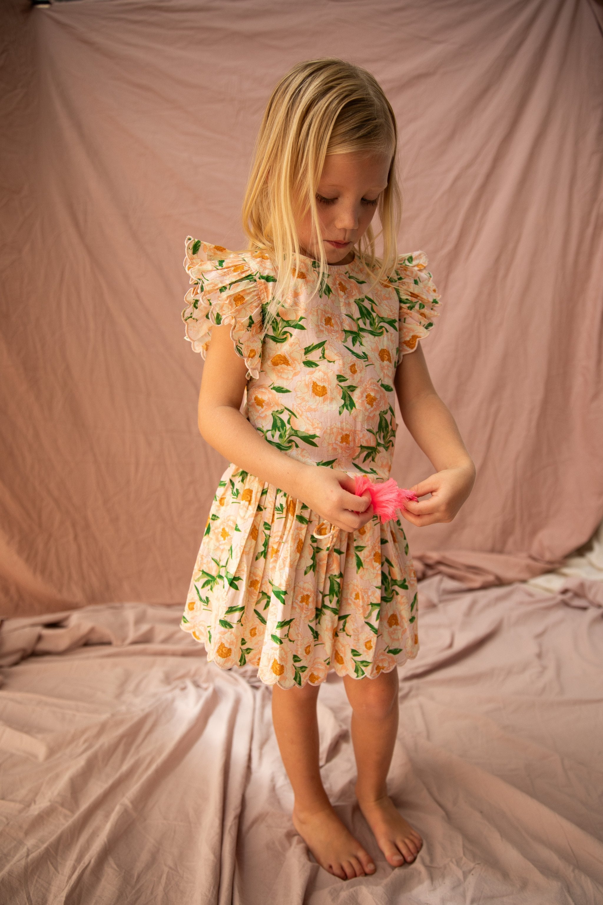 A young girl with shoulder-length blonde hair stands barefoot on a pink fabric backdrop. She wears the BELLA + LACE Vivian Top Soirée Bouquet/Strawberry Sundae, characterized by its flowery pattern, ruffled sleeves, and delicate scalloped embroidery. She holds a small pink object and looks down at it intently.