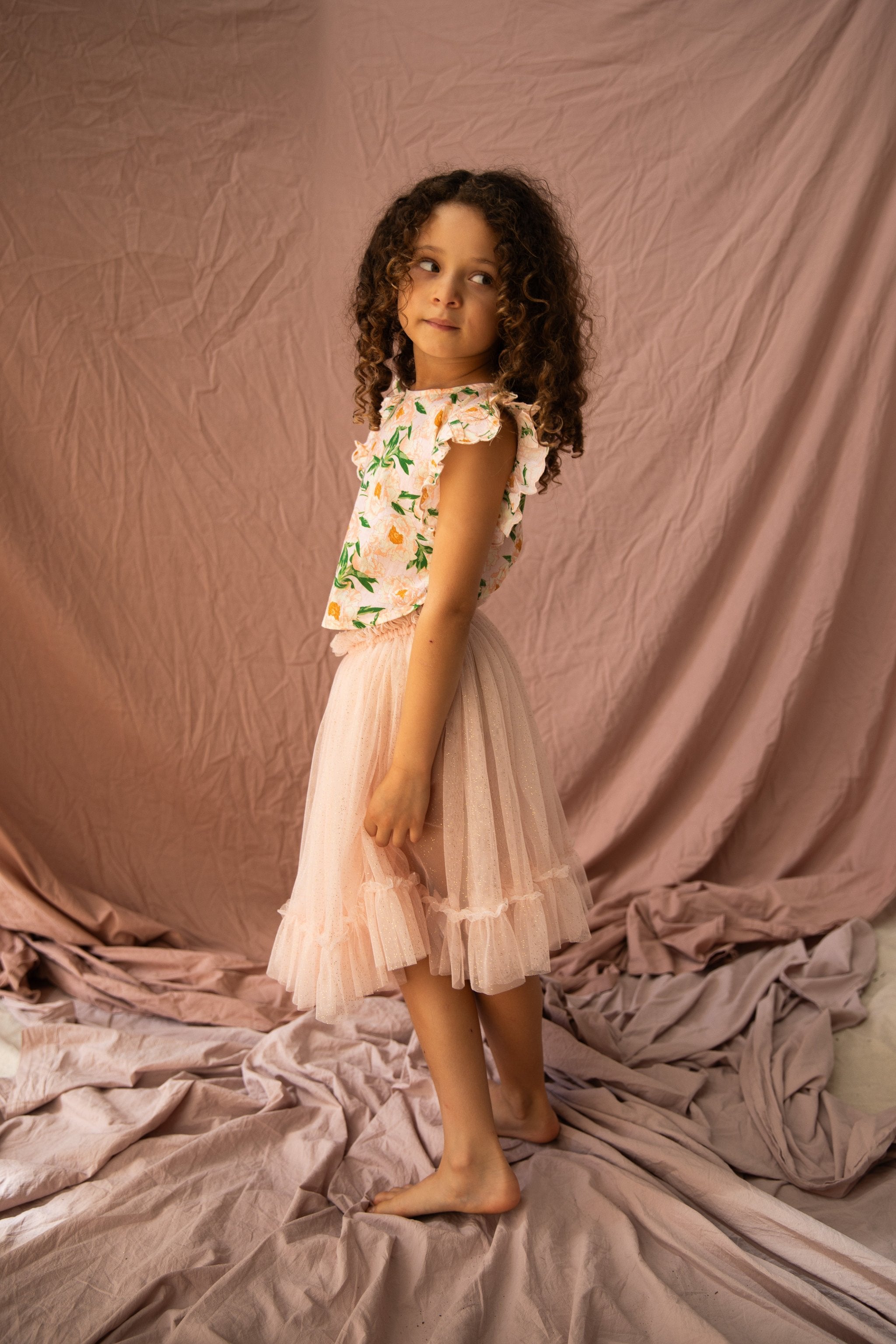 A young girl with curly hair stands barefoot on a soft, crumpled fabric backdrop, wearing the Vivian Top Soirée Bouquet/Strawberry Sundae by BELLA + LACE and a pink tulle skirt. The light cotton voile of her floral top adds to the airy elegance. She looks off to the side with a thoughtful expression, while the draped pink fabric complements her skirt perfectly.