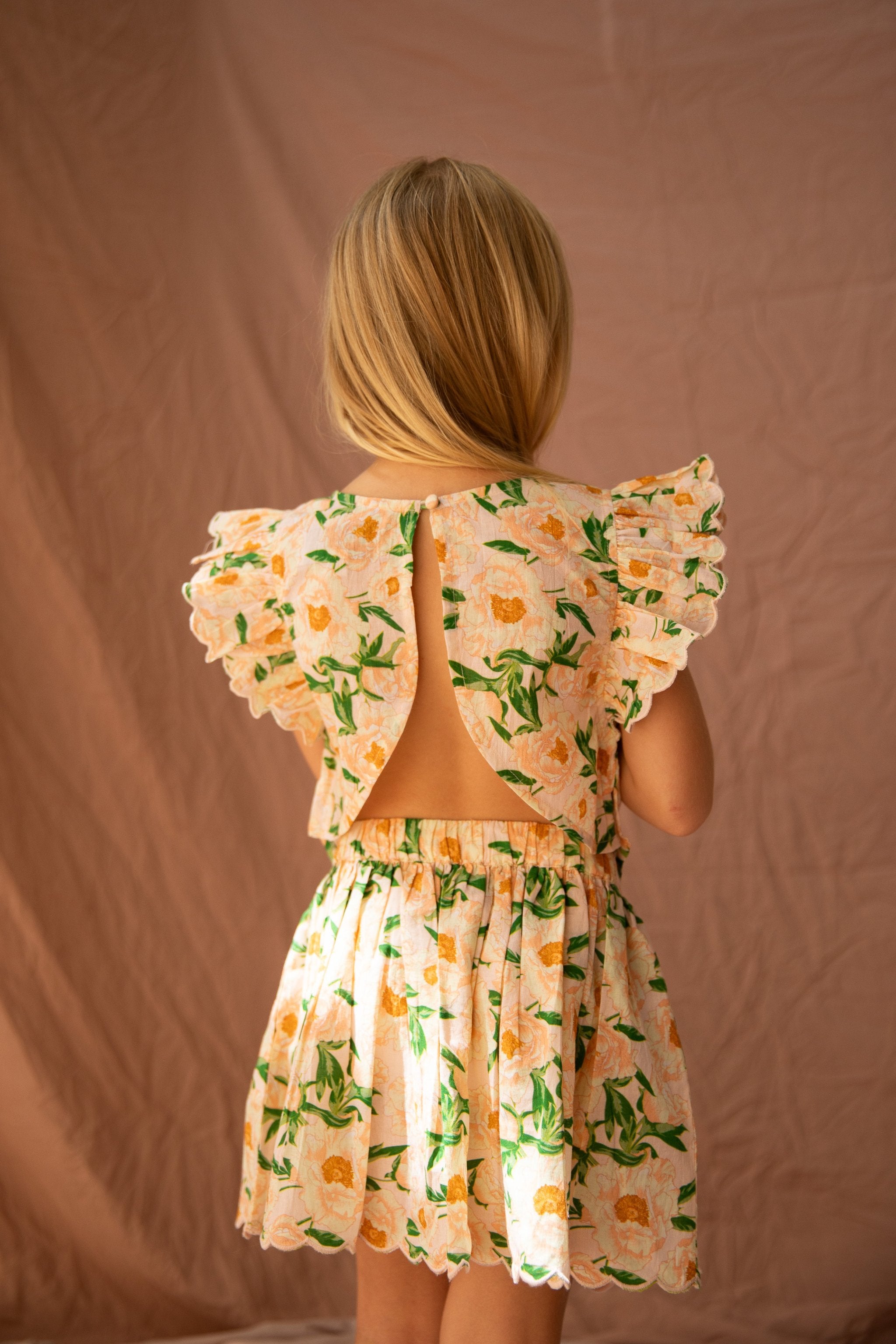 A young girl with long blonde hair stands facing away from the camera, wearing the Vivian Top in Soirée Bouquet/Strawberry Sundae by BELLA + LACE. The light cotton voile top features a pattern of peach-colored flowers with green leaves, set against a beige background, and boasts ruffled sleeves and an open-back design.