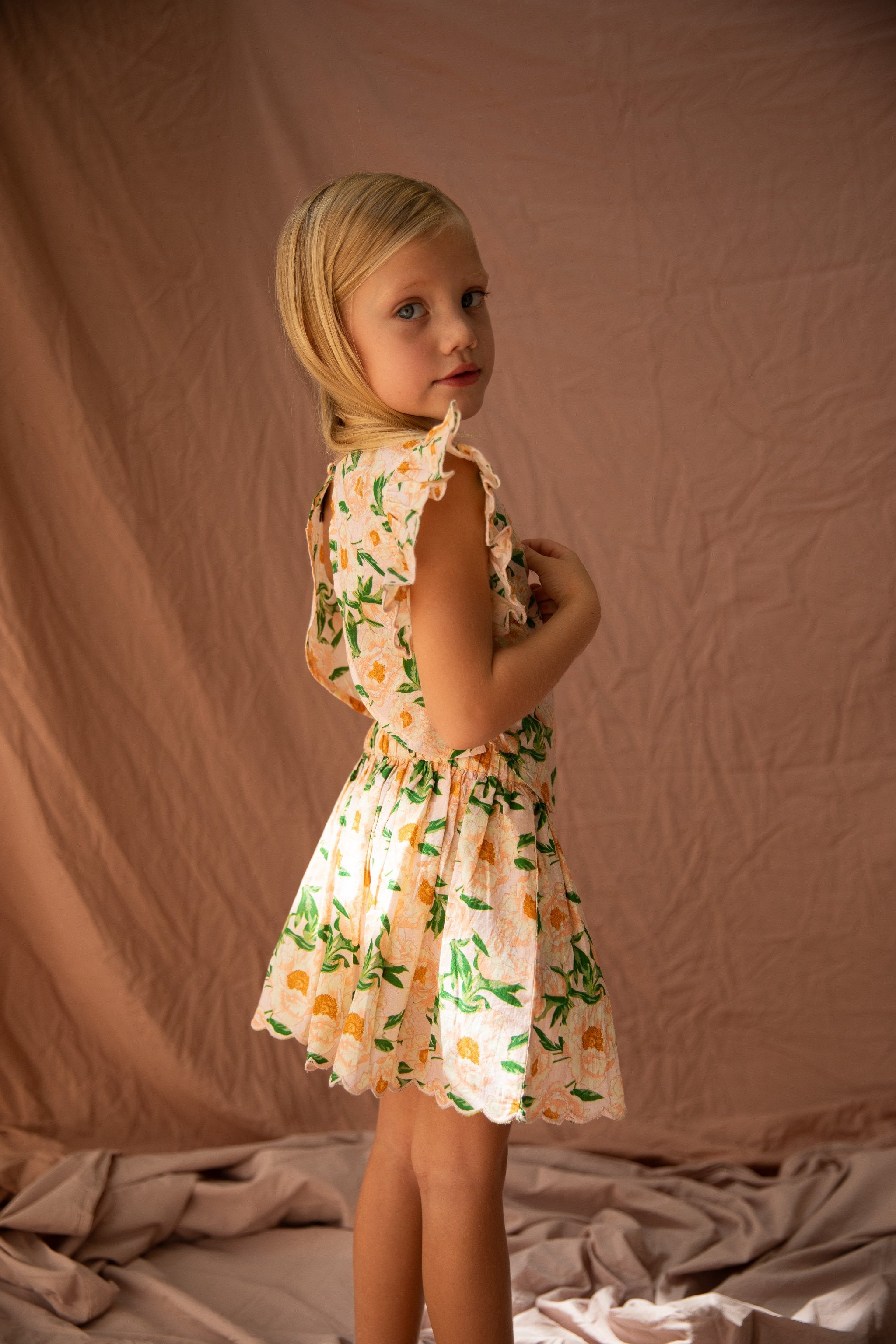 A young girl with blonde hair stands in a light-colored room with a fabric backdrop. She is wearing the Vivian Top Soirée Bouquet/Strawberry Sundae by BELLA + LACE, a knee-length floral-patterned dress made of light cotton voile, featuring ruffled sleeves and scalloped embroidery. She gazes over her shoulder with a subtle, calm expression under soft, warm lighting.