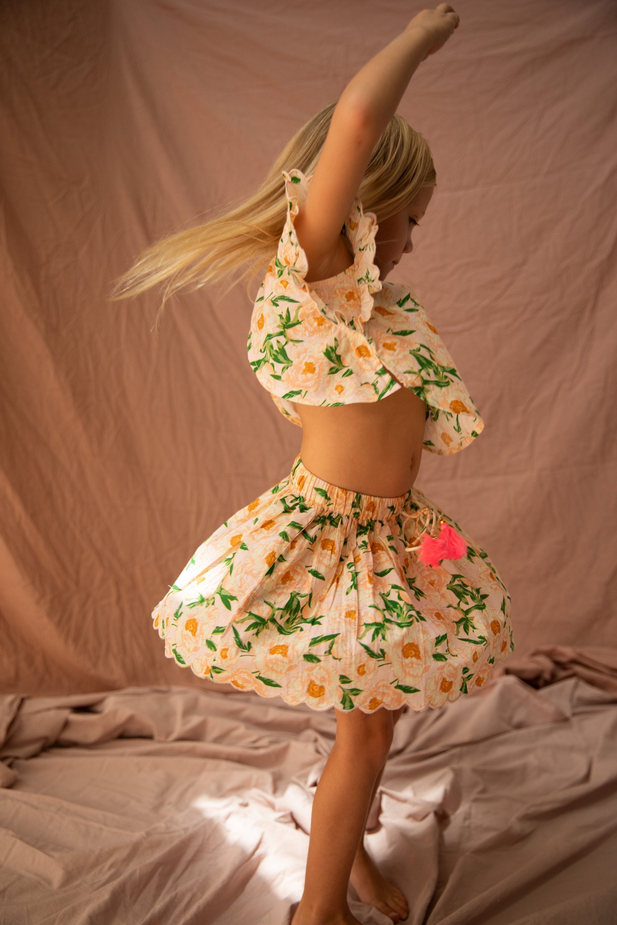 A young girl with blonde hair twirls in the BELLA + LACE Vivian Top Soirée Bouquet/Strawberry Sundae, made of light cotton voile and featuring an off-the-shoulder ruffled top with a matching skirt. The pink flower pattern adds a touch of charm against the beige and peach fabric background.