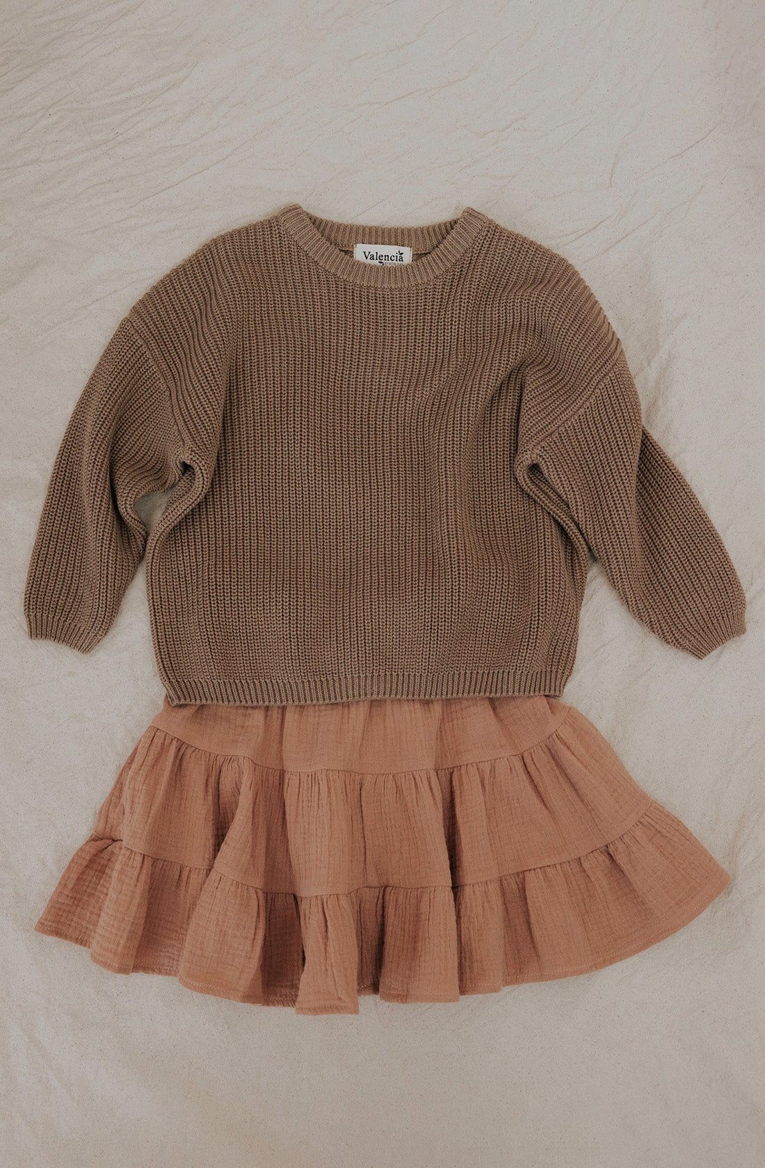 A Farm Knit Cinnamon from VALENCIA BYRON BAY, a neutral cinnamon-colored, long-sleeve, knitted cotton sweater with an oversized fit, is paired with a light brown, tiered ruffled skirt and laid out on a cream-colored fabric.