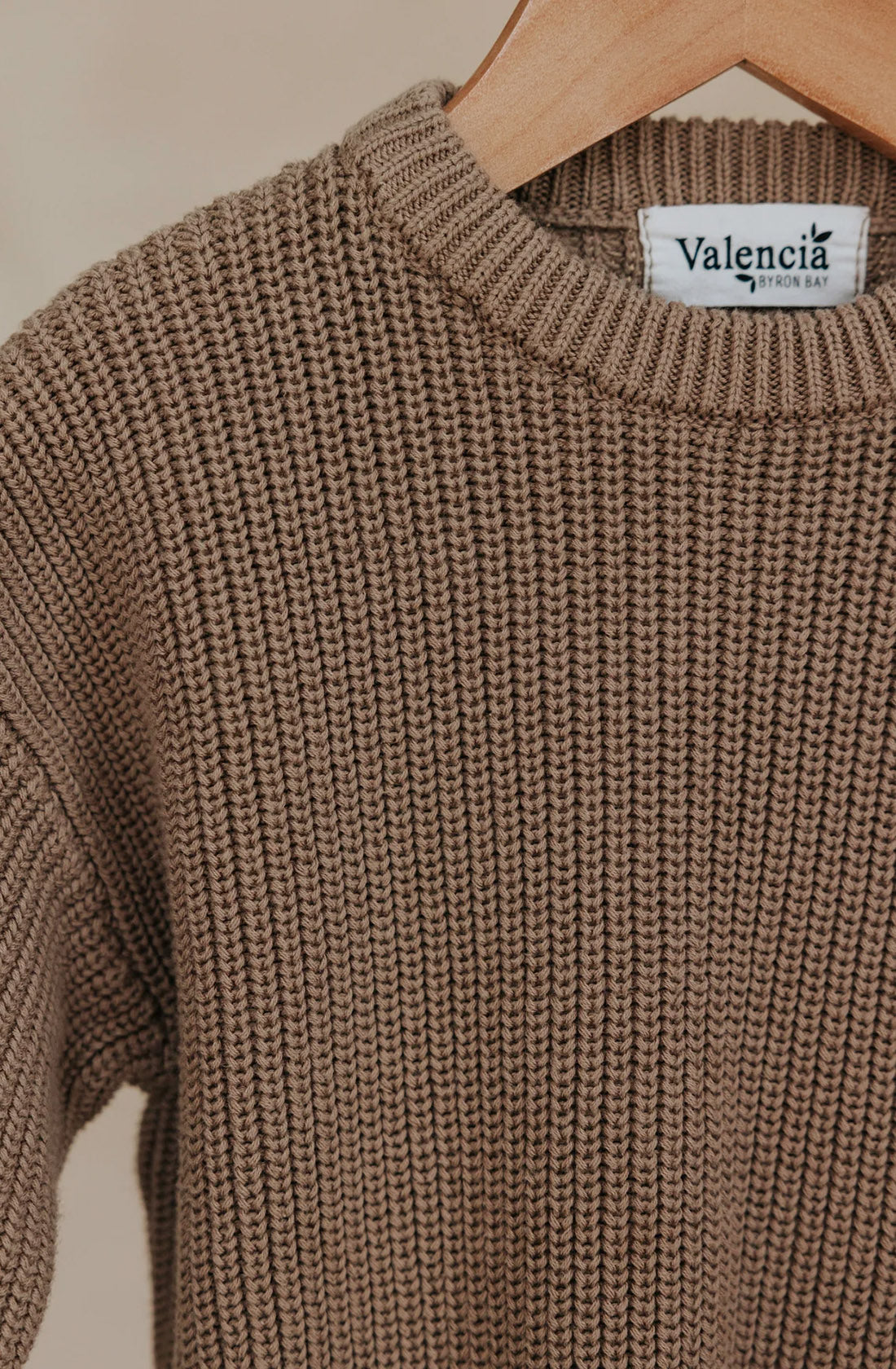 A close-up image of the Farm Knit Cinnamon, a brown, ribbed knit sweater in a neutral cinnamon color on a wooden hanger. This thick, cozy cotton sweater has an oversized fit and features a tag near the collar that reads "VALENCIA BYRON BAY." The textured knit pattern adds to its cozy appeal.