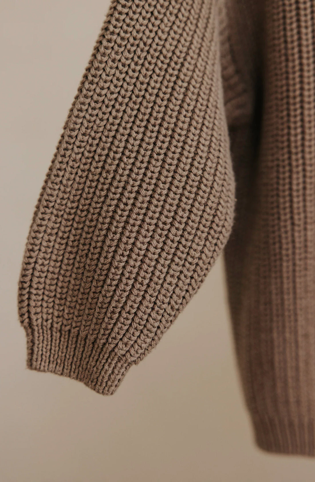 Close-up of a Farm Knit Cinnamon cotton sweater sleeve by VALENCIA BYRON BAY. The sleeve features a ribbed texture with dense, chunky stitching and ends in a small cuff. The material appears thick and cozy, making it ideal for cold weather. The background is uncluttered.