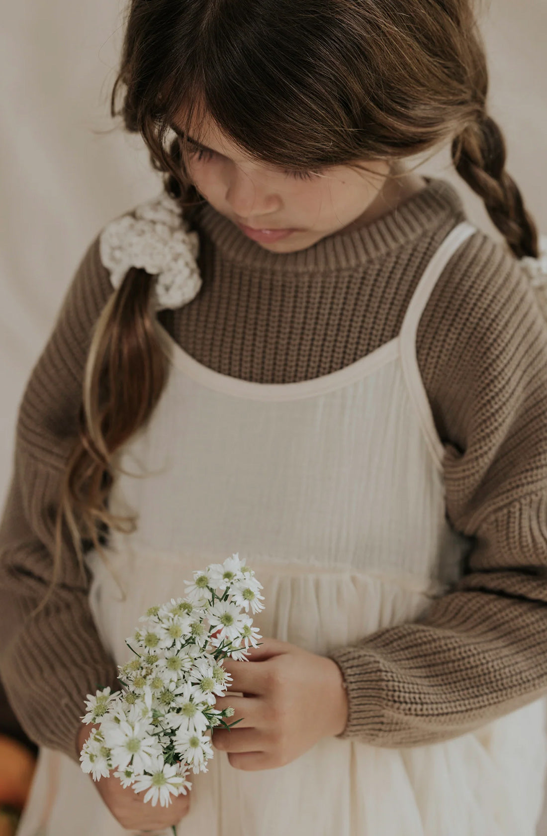 A young girl with braided hair, wearing the VALENCIA BYRON BAY Farm Knit Cinnamon oversized cotton sweater over a white dress, looks down at a small bouquet of white flowers she is holding in her hands. The background is softly blurred, enhancing the gentle and contemplative moment.