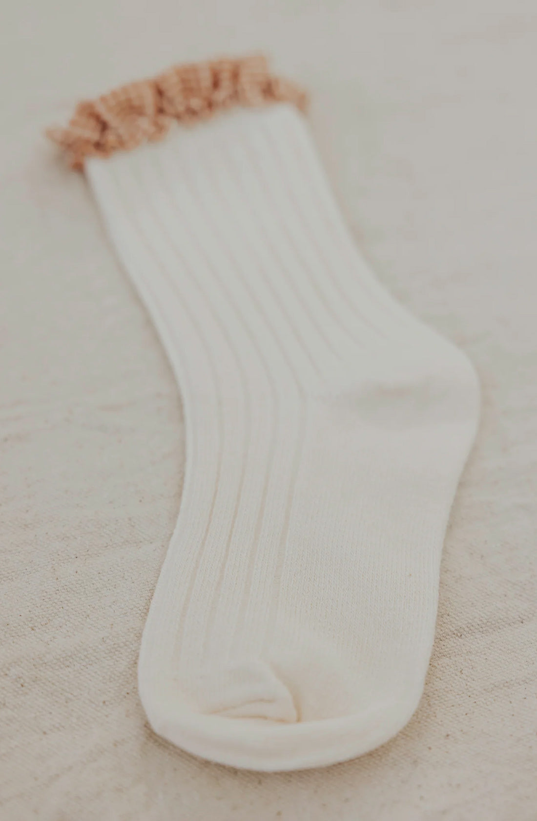 Lying flat on a textured, light-colored surface is a single Frilly Socks Milk by VALENCIA BYRON BAY, featuring ribbed detailing and a frilly peach-colored cuff in pristine white cotton.