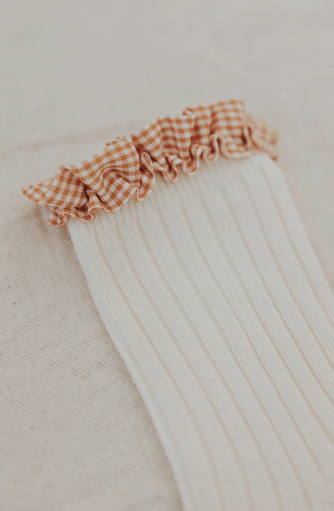 A close-up image of a white ribbed fabric with a ruffled edge displaying a brown and white gingham checkered pattern. The fabric appears soft and textured, reminiscent of the Frilly Socks Milk from VALENCIA BYRON BAY's clothing collection, likely set against a neutral background.