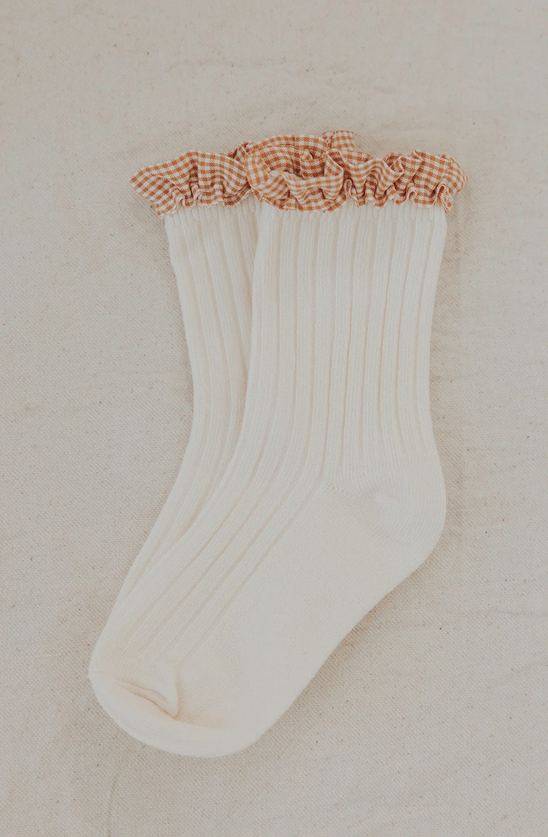 A pair of Frilly Socks Milk by VALENCIA BYRON BAY, featuring white ribbed cotton and an orange and white gingham ruffle at the top, laid flat on a light beige fabric surface. These charming socks add a delightful touch to any outfit.