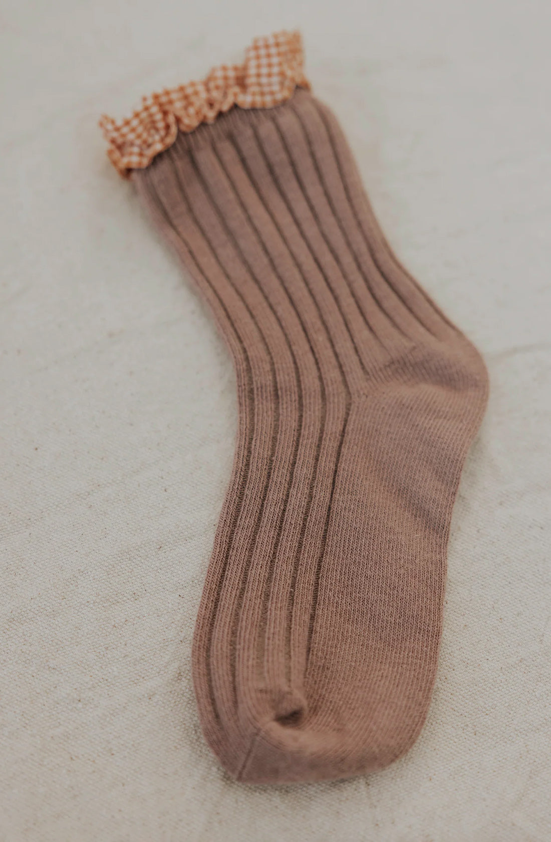 The Frilly Socks Nutmeg by VALENCIA BYRON BAY, a single brown ribbed sock with a ruffled checkered cuff, perfect for protecting little feet, is laid flat on a light, textured surface.