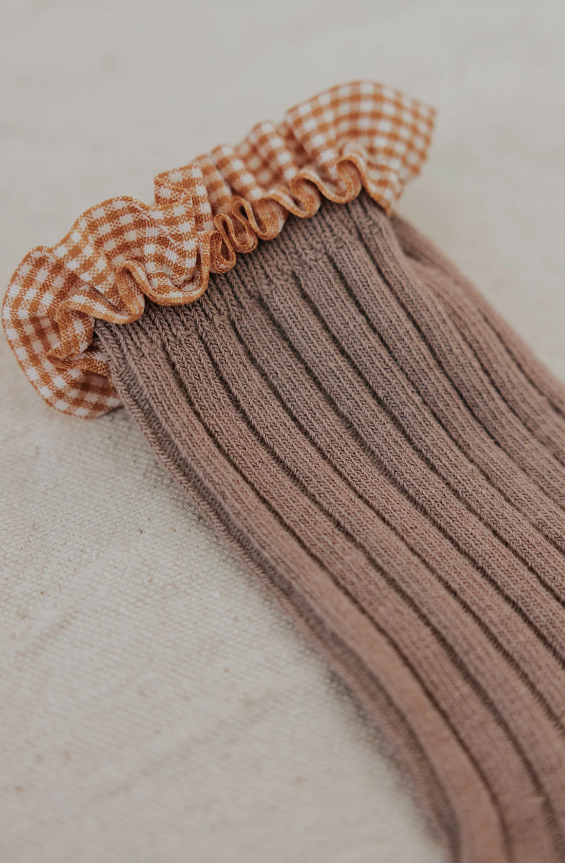 Close-up of a ribbed Frilly Socks Nutmeg by VALENCIA BYRON BAY in brown cotton with a ruffled gingham top in white and orange. The sock is laid out flat on a neutral, textured surface, perfect for protecting little feet during colder months.