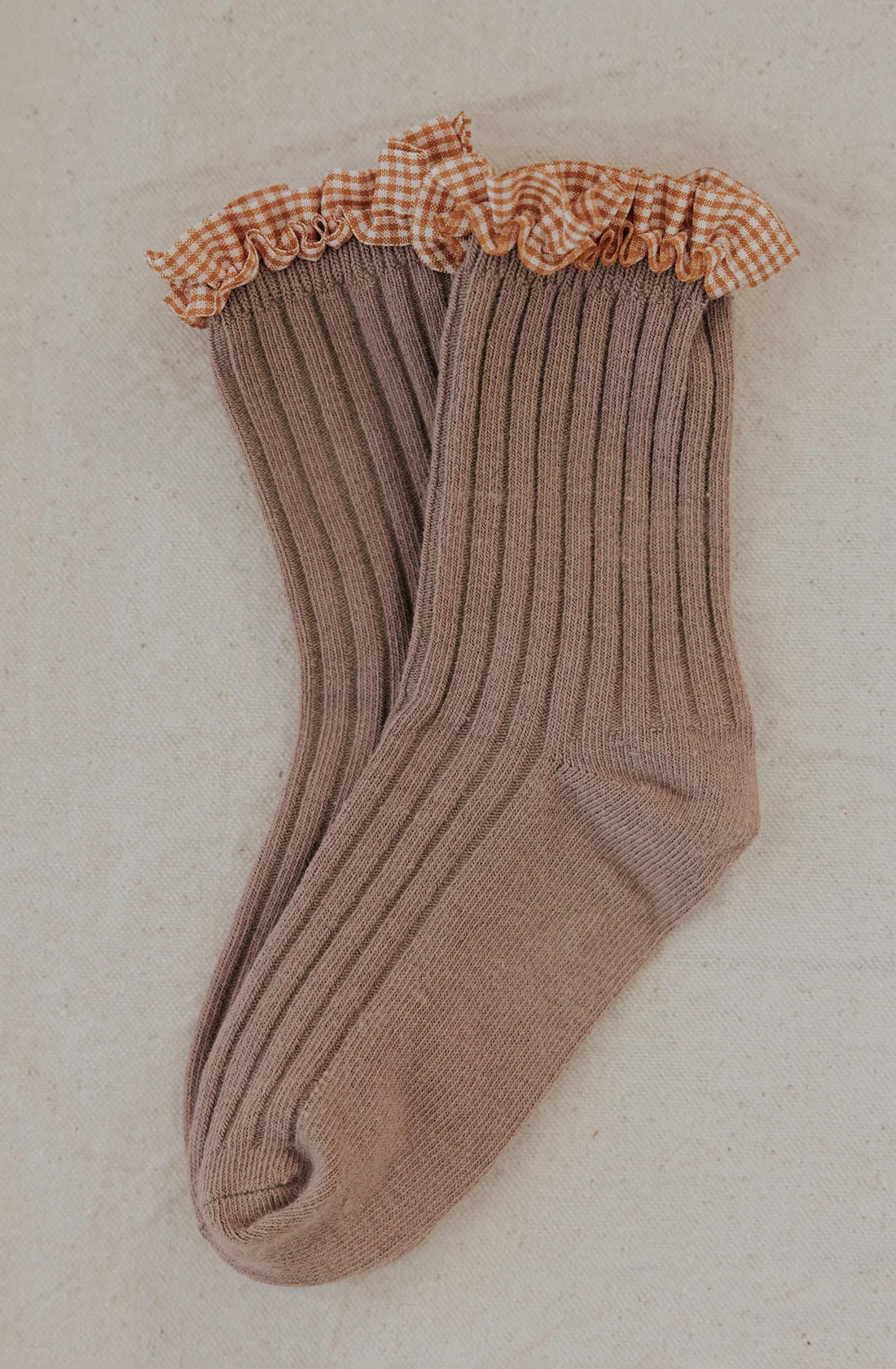 VALENCIA BYRON BAY's Frilly Socks Nutmeg are a pair of ribbed beige cotton socks with a ruffled top edge featuring a beige and white gingham pattern, perfect for keeping little feet protected on a light, neutral-colored fabric background.