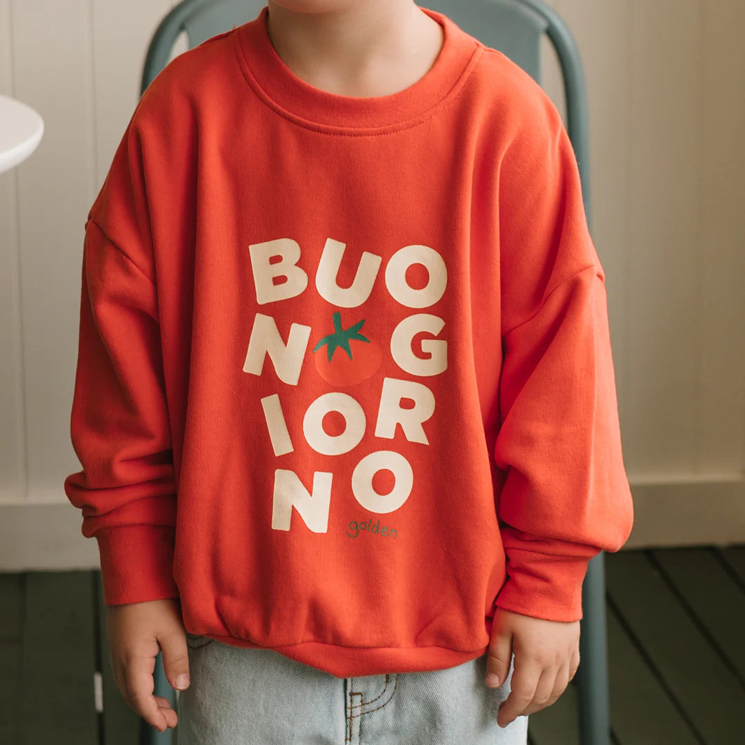 A child wears the GOLDEN CHILDREN Buongiorno Sweater Napoli—a relaxed fit, soft french terry fleece cotton sweater with “BUONGIORNO” and a tomato graphic in white on the front—while standing indoors.