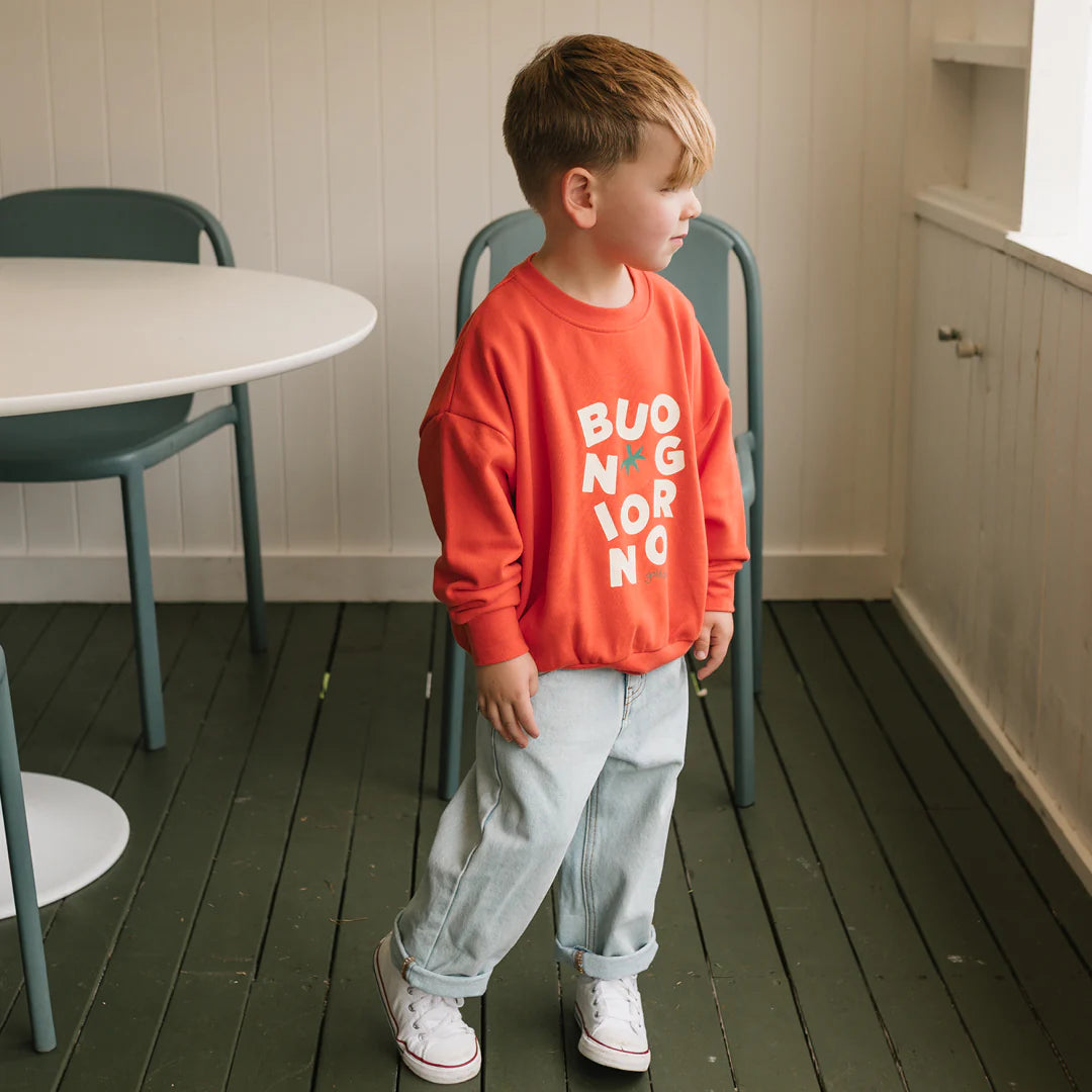 A young boy wearing the GOLDEN CHILDREN - Buongiorno Sweater Napoli in a relaxed fit, with light blue jeans and white sneakers, stands indoors beside a green chair and white table on a wooden floor, looking to the right.