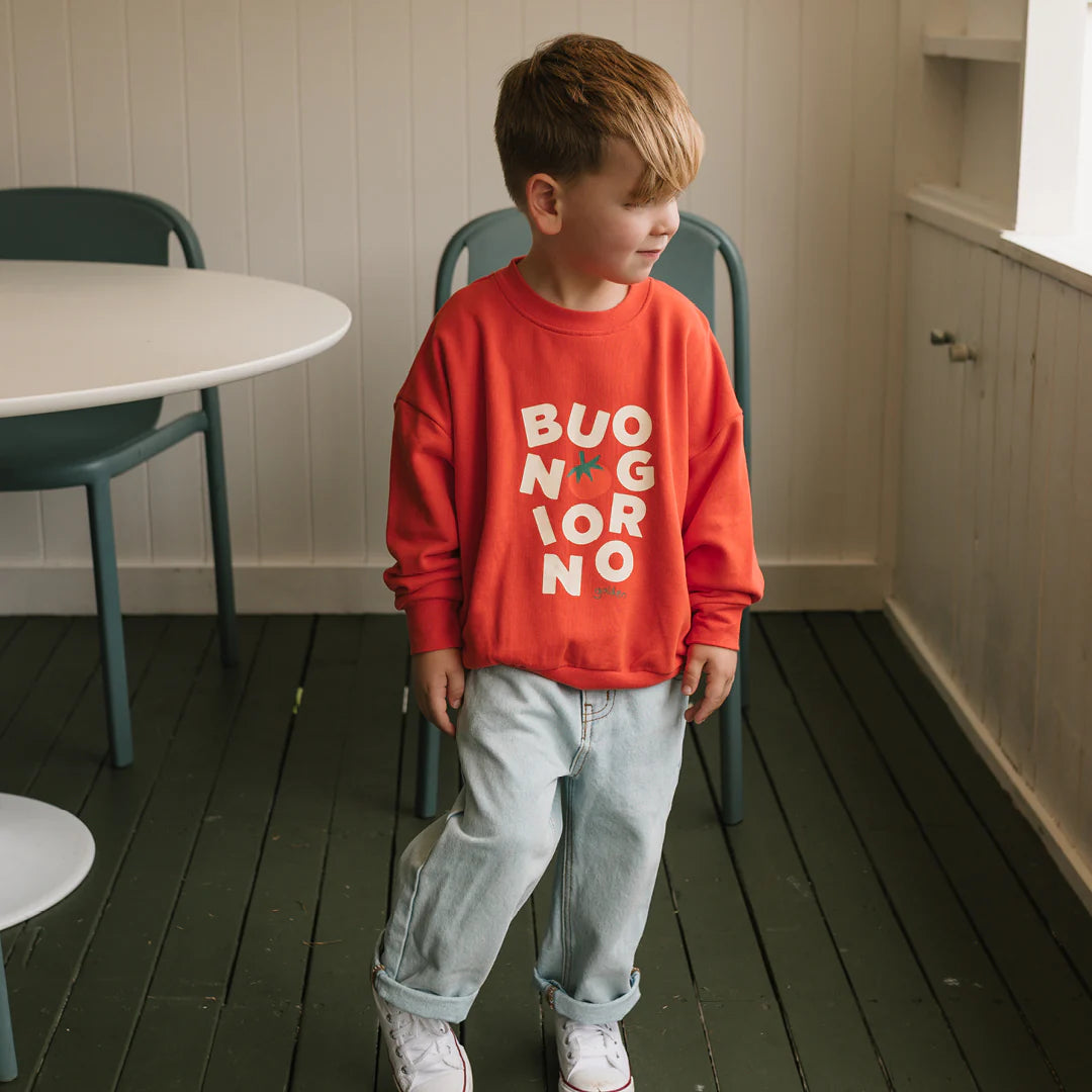 A young child with light brown hair wears the GOLDEN CHILDREN Golden Children - Buongiorno Sweater Napoli in bright red, paired with light blue jeans and white sneakers, standing indoors near a white table and green chair.