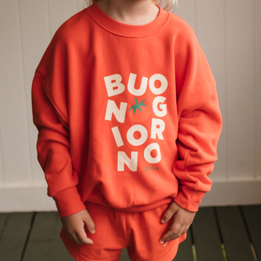 A child models the GOLDEN CHILDREN Buongiorno Sweater Napoli—a relaxed fit, orange cotton french terry fleece with “BUONGIORNO” in white and a tomato for the “O.” Child’s face is not visible.