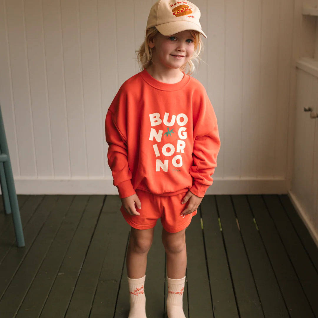 A young child smiles brightly indoors, wearing the GOLDEN CHILDREN - Buongiorno Sweater Napoli and shorts in soft french terry fleece cotton, paired with a beige cap and cream socks on green wooden flooring.