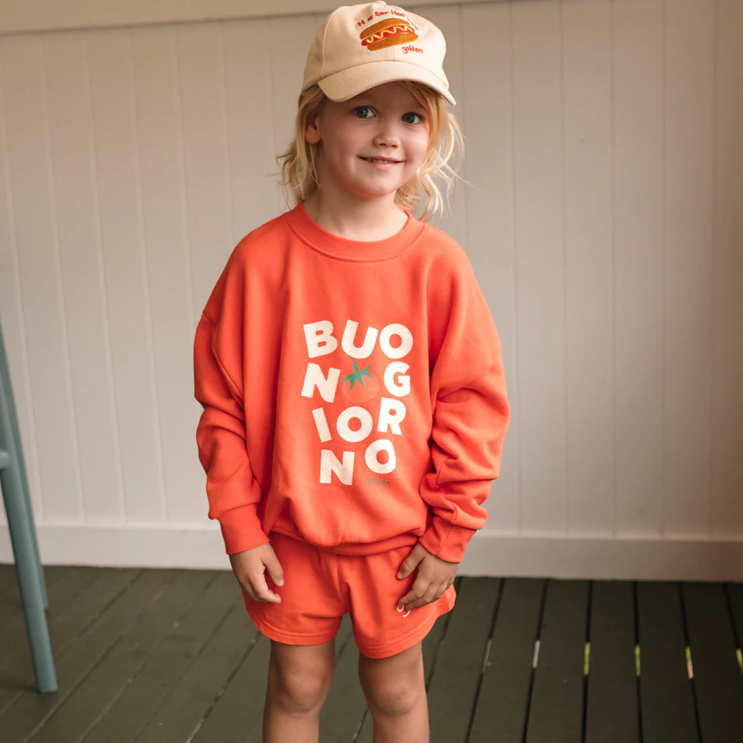 A young child in a relaxed fit GOLDEN CHILDREN Buongiorno Sweater Napoli, orange shorts, and a beige cap with a hot dog design stands indoors on a green wooden floor.