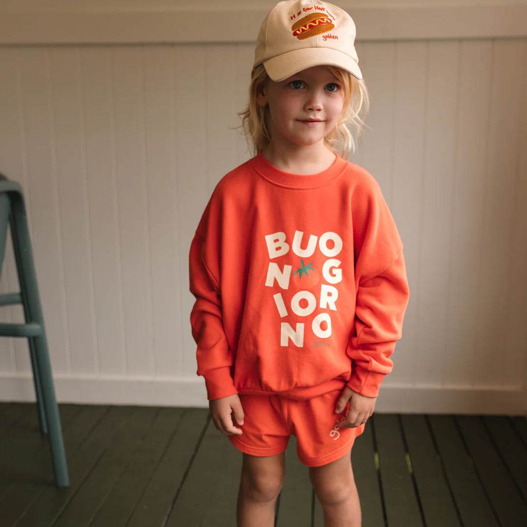 A young child wears the GOLDEN CHILDREN Golden Children - Buongiorno Sweater Napoli in orange French terry fleece cotton, matching shorts, and a beige hot dog graphic baseball cap, standing indoors on a wooden floor.