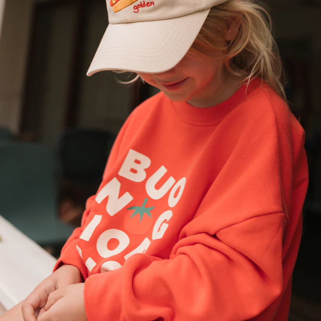 A child wears the relaxed fit GOLDEN CHILDREN Buongiorno Sweater Napoli in orange, featuring bold white letters and a tomato graphic, paired with a beige cap, smiling softly with an out-of-focus background.