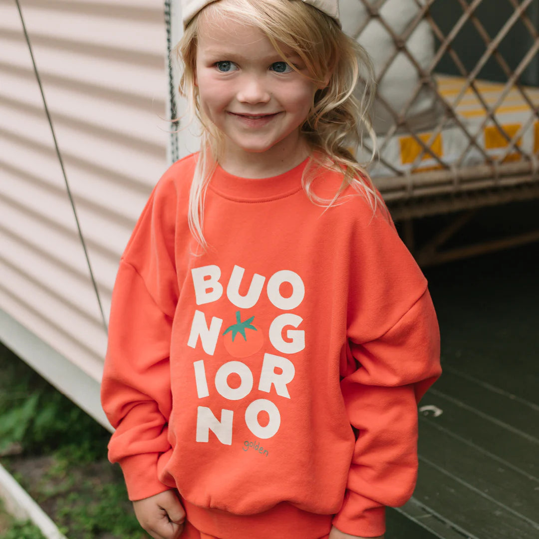 A smiling young child with long blonde hair wears the GOLDEN CHILDREN Buongiorno Sweater Napoli, a relaxed-fit french terry fleece cotton sweater featuring "BUONGIORNO" and a tomato graphic, standing by striped building siding.