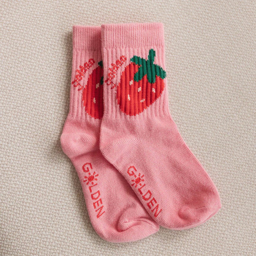 Golden Children - Strawberry Cake Socks Strawberry by GOLDEN CHILDREN are kids’ cotton socks with a large red strawberry and green leaves, plus "GOLDEN" in red near the toes, shown on a textured beige surface.
