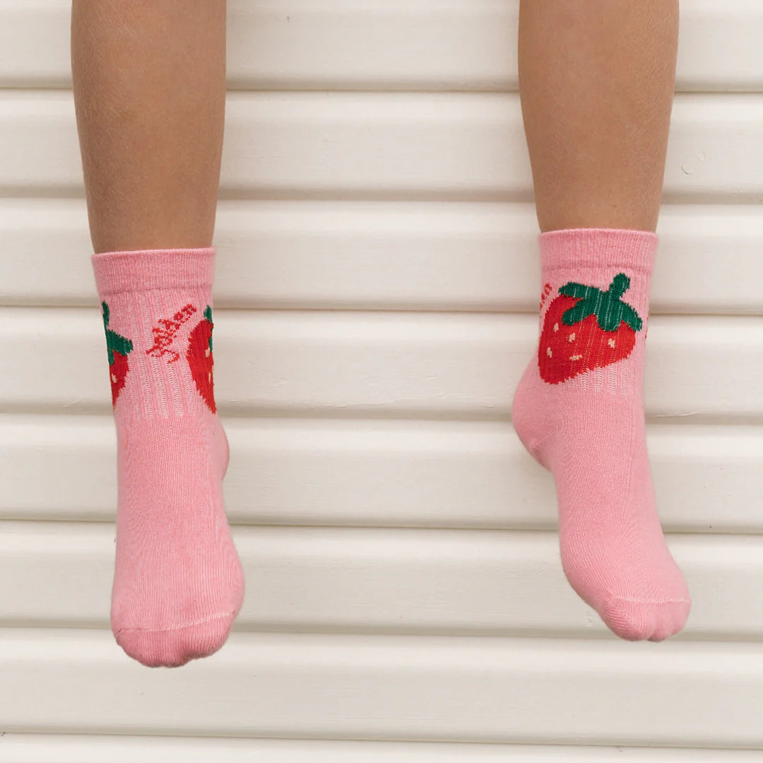 A child’s legs in GOLDEN CHILDREN Golden Children - Strawberry Cake Socks Strawberry—kids’ cotton socks with red strawberries—rest on a crisp white, ridged background.