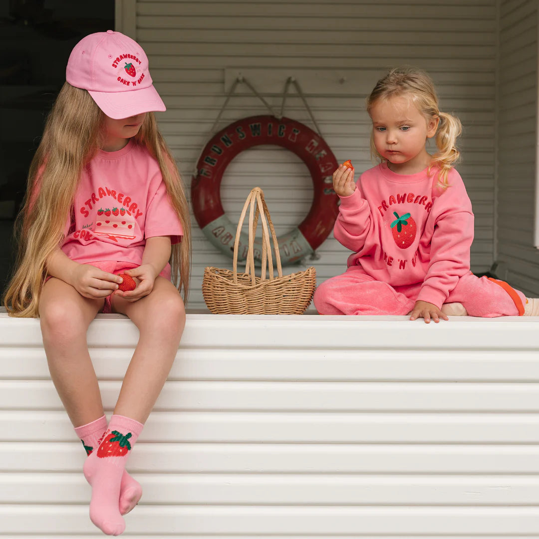 Two young girls sit on a white porch in pink, strawberry-print outfits. One wears GOLDEN CHILDREN’s Golden Children - Strawberry Cake Socks Strawberry and a cap; the other is barefoot. Both hold strawberries, with wicker and life preserver nearby.