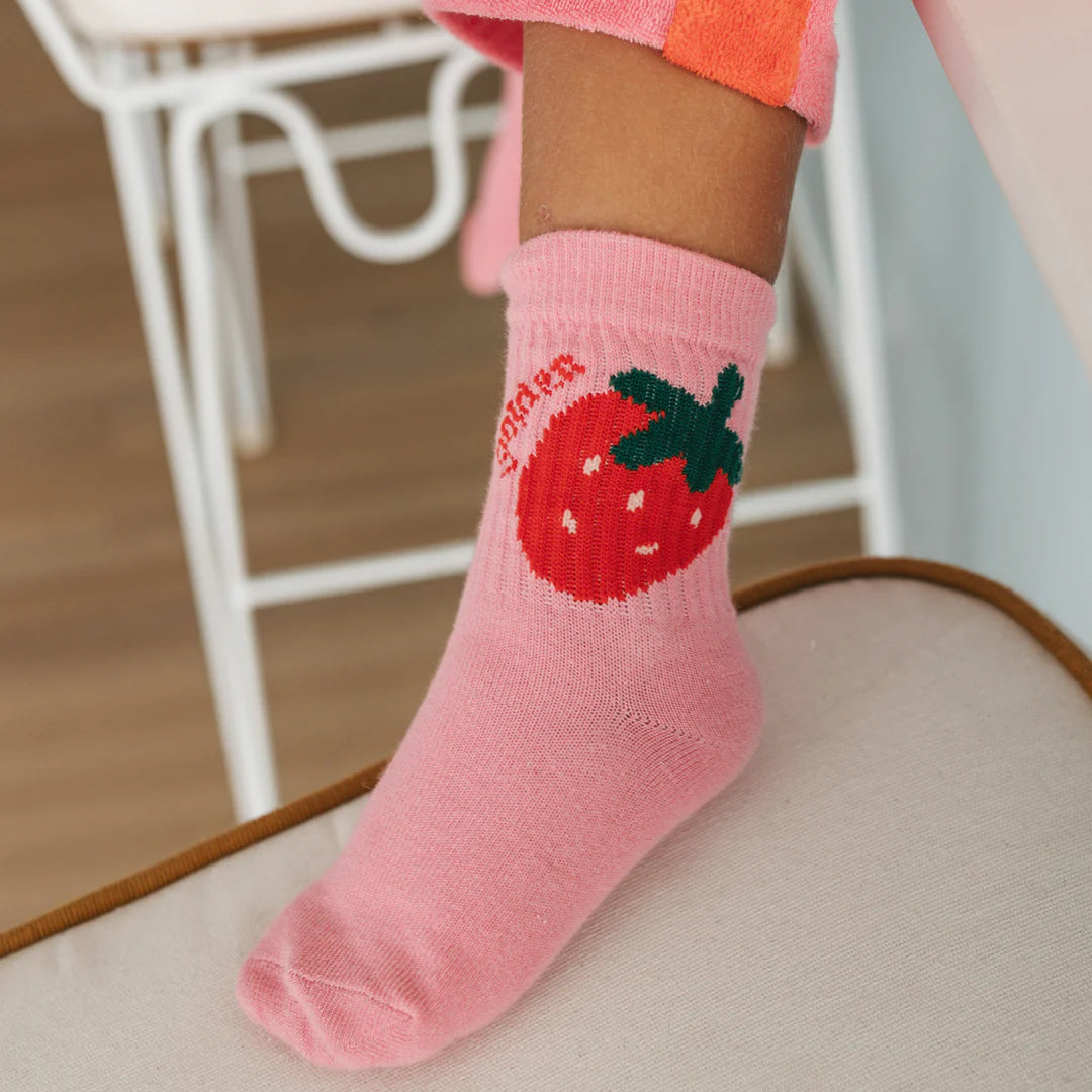 A child wears GOLDEN CHILDREN's Golden Children - Strawberry Cake Socks Strawberry—pink cotton socks with a red strawberry and green leaves, plus "strawberry" text. The child sits on a light cushion near a white chair.
