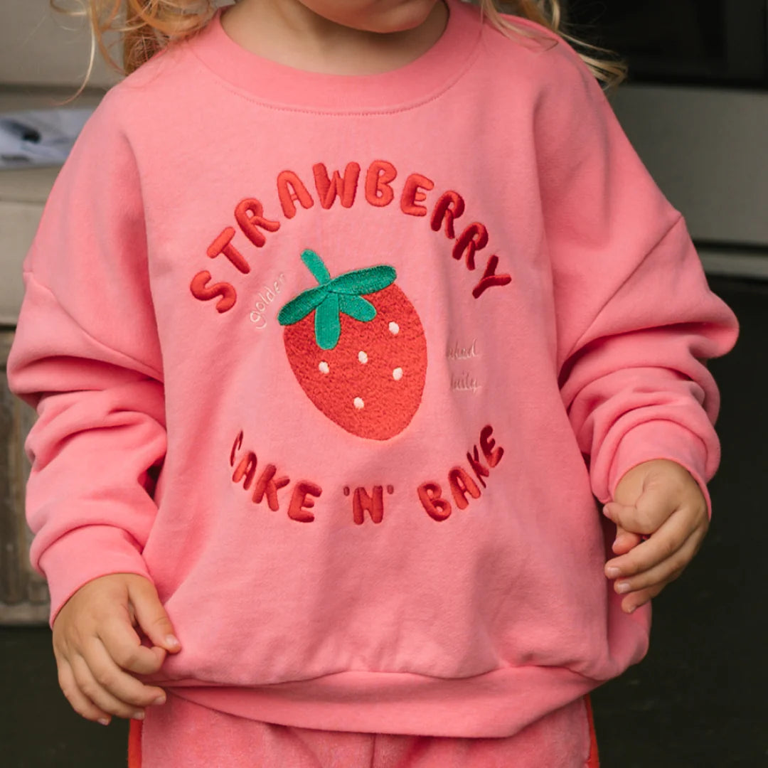 A child wears the GOLDEN CHILDREN Strawberry Cake Sweater Meringue Pink, made from french terry fleece cotton, with a large embroidered strawberry and "STRAWBERRY CAKE 'N' BAKE" on the front. The child's face is not shown.