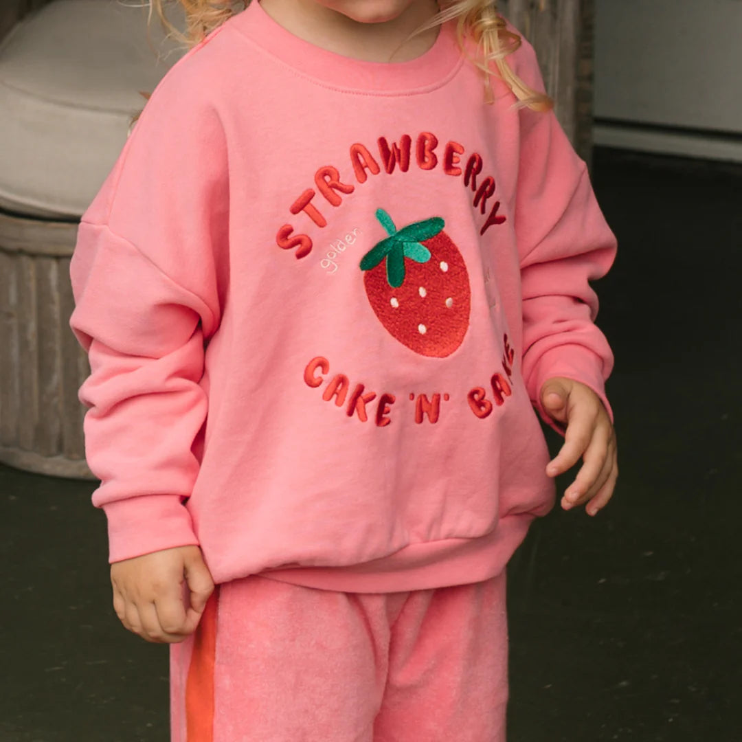 A young child in the GOLDEN CHILDREN Golden Children - Strawberry Cake Sweater Meringue Pink, a relaxed-fit french terry fleece with embroidered strawberry and “Strawberry Cake 'N' Bake” on front, paired with matching pink pants. Face partially out of frame.