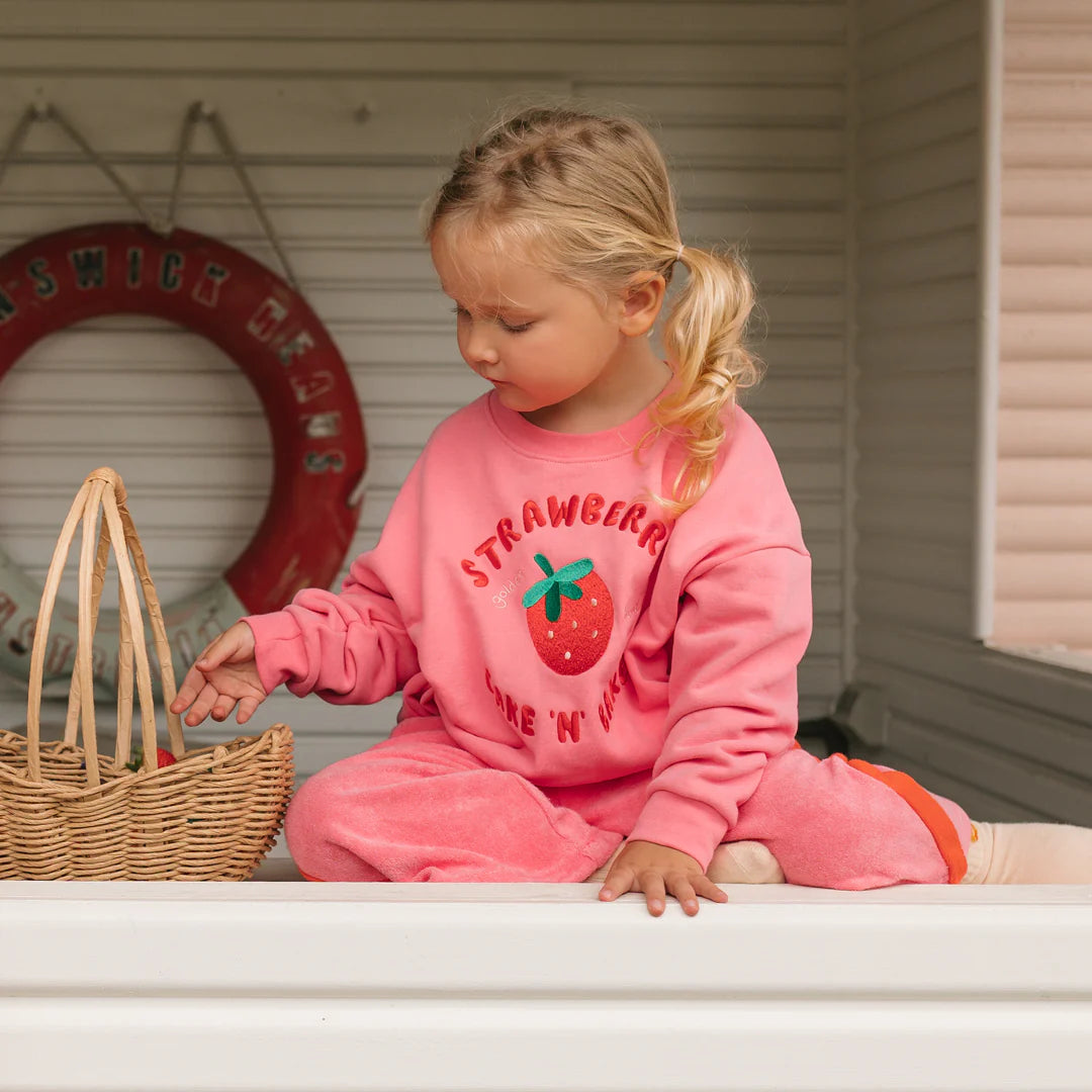 A young child in the GOLDEN CHILDREN Golden Children - Strawberry Cake Sweater Meringue Pink, made from french terry fleece cotton, sits cross-legged on a white surface, reaching into a wicker basket with a life preserver in the background.